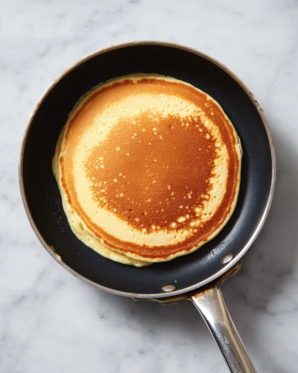 A single round pancake with a light golden brown center and slightly lighter edges sits in a black frying pan with a silver handle. The pancake fills most of the pan, showing a soft and fluffy texture with some small bubbles on the surface. The pan rests on a white marbled surface, and no other objects or food items are visible in the scene. photo taken with an iphone --ar 4:5 --v 7