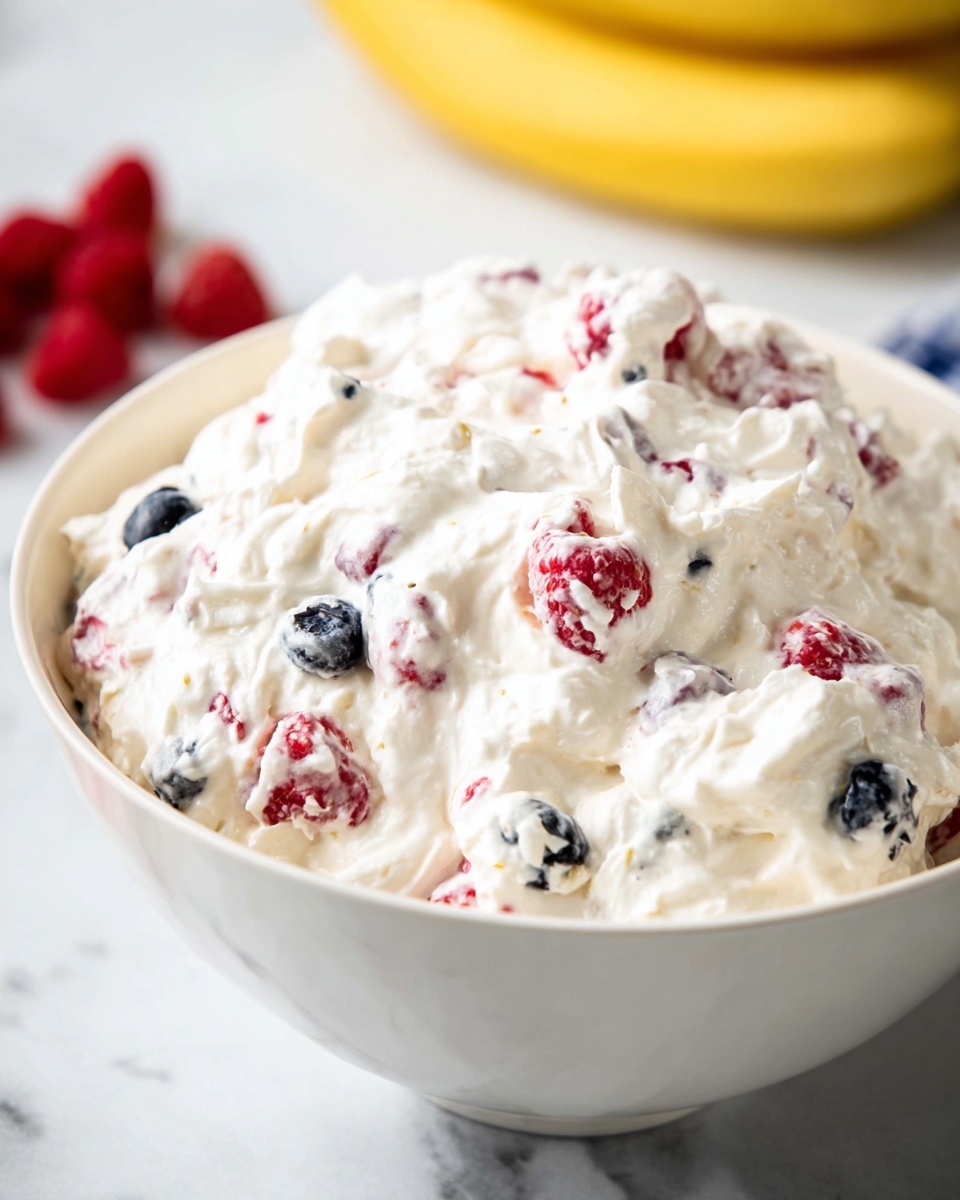 A large white bowl is filled with a creamy, thick white mixture, studded with red raspberries and dark blue blueberries, creating a textured and fluffy appearance. The berries are mixed evenly throughout the whipped white cream, showing bits of red and blue beneath the soft peaks of the cream. The bowl sits on a white marbled surface, and in the soft background, a yellow banana and some red raspberries are visible but out of focus. The overall look is fresh and light, with a mix of smooth and bumpy textures from the whipped cream and fruit. photo taken with an iphone --ar 4:5 --v 7