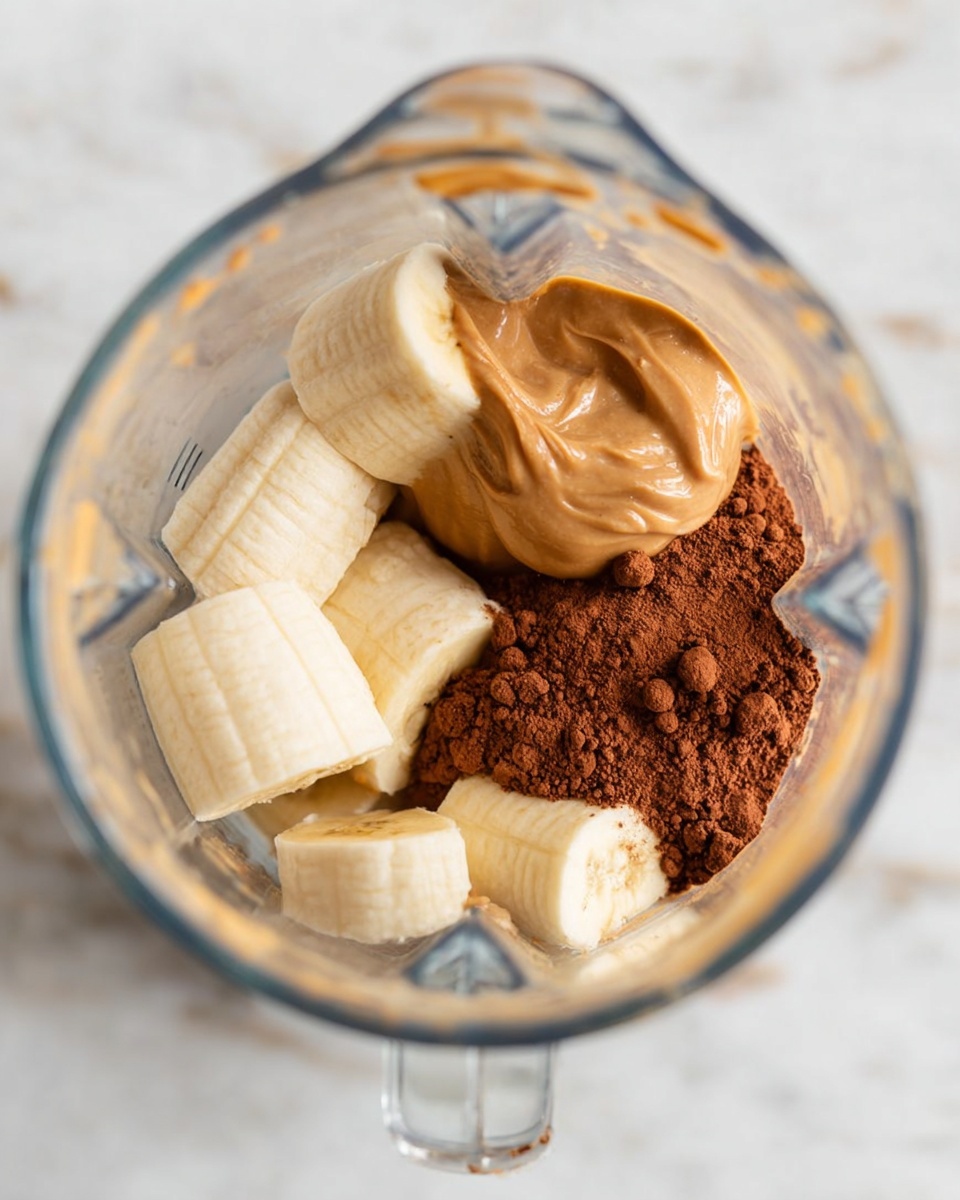 Inside a clear blender jar, there are three main ingredients in separate layers: at the bottom, large pieces of pale yellow frozen banana with a smooth, frosty texture; next to the banana, a pile of rich brown cocoa powder with a dry and powdery look; on top of the banana and cocoa, a thick dollop of light brown creamy peanut butter with a smooth, slightly swirled surface. The jar sits on a white marbled surface. photo taken with an iphone --ar 4:5 --v 7