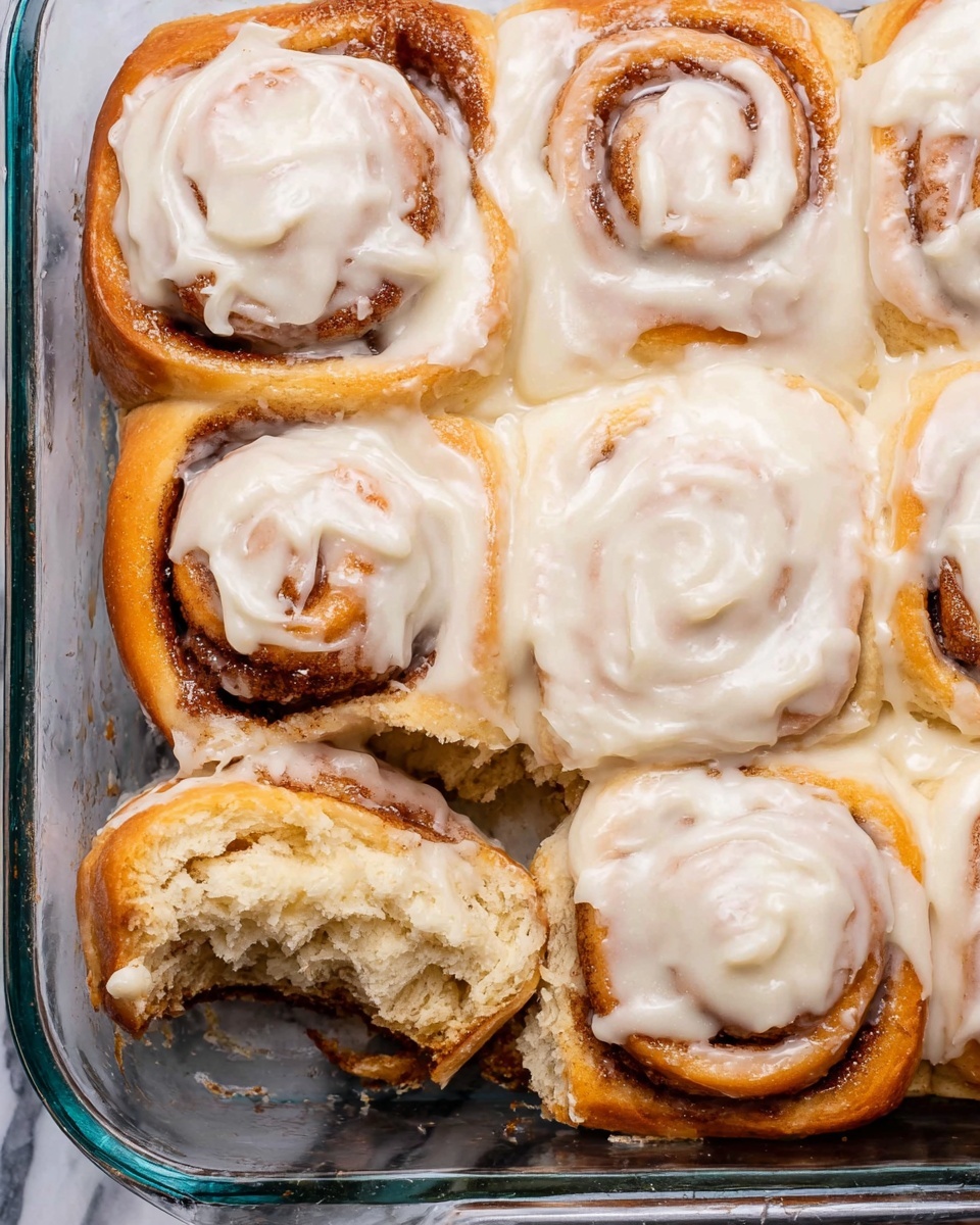 A soft, thick cinnamon roll sits on a white plate with a silver fork resting in front of it. The cinnamon roll has three visible layers: the bottom layer is a pale, fluffy dough, the middle layer is a rich cinnamon swirl with a dark brown, slightly glossy texture, and the top layer is covered with creamy, white icing that looks smooth and slightly melting, dripping down the sides. The roll’s torn edges show soft, airy inside dough mixed with cinnamon. The plate is set on a white marbled surface. Photo taken with an iphone --ar 4:5 --v 7