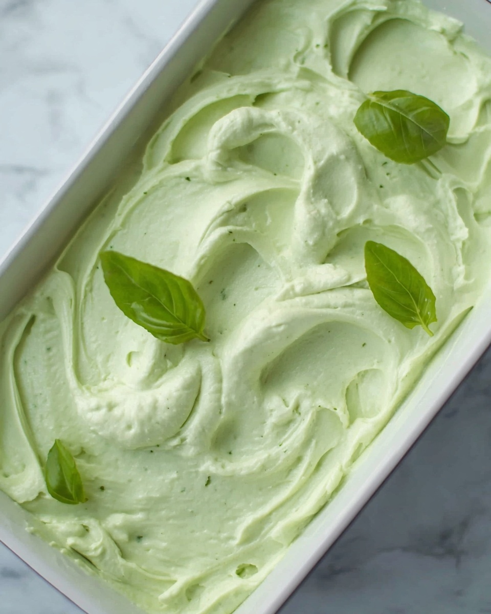 The image shows a close-up of a creamy, light green mixture spread in a rectangular container. The texture is smooth with gentle swirls and soft peaks on top. There are a few bright green basil leaves placed on the surface for decoration. The container is white and is set against a white marbled surface. photo taken with an iphone --ar 4:5 --v 7