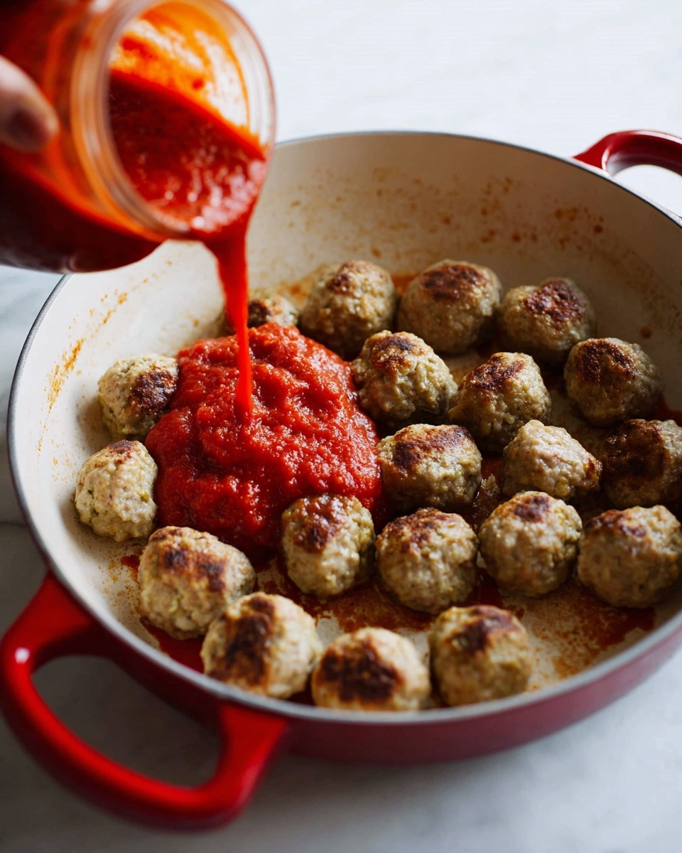 A white round pan with red handles holds about 20 light brown meatballs with a slightly crispy texture on a white marbled surface. A woman's hand pours thick, bright red tomato sauce over the meatballs from a jar, creating a contrast between the smooth, chunky sauce and the browned meatballs. The pan’s inside surface shows some marks from cooking, adding to the homemade look. photo taken with an iphone --ar 4:5 --v 7