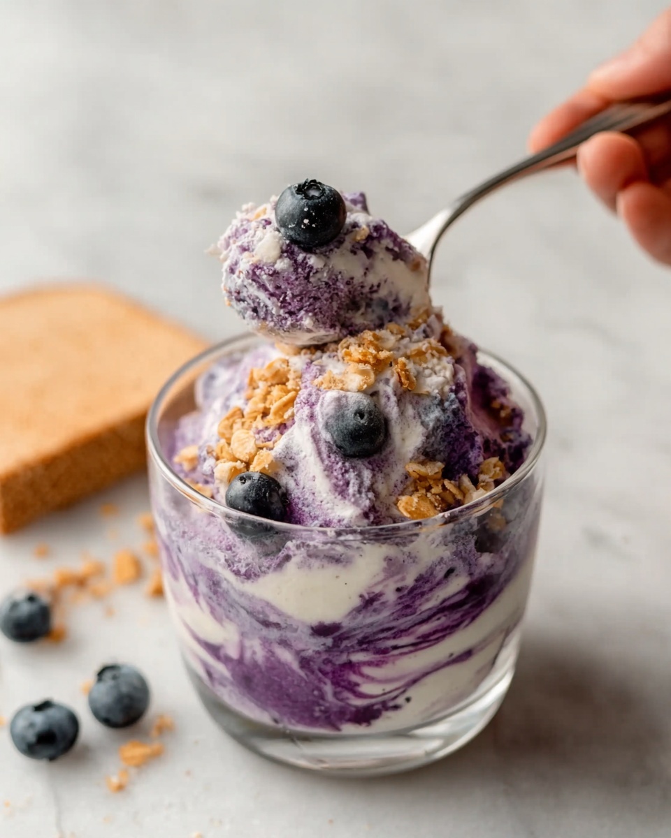 A clear glass bowl filled with three layers of purple and white swirled creamy dessert, topped with three whole fresh blueberries and sprinkled with small golden brown granola pieces. A silver spoon is inside the bowl, scooping a portion of the dessert, held by a woman's hand from the right side. The background shows a white marbled surface with a corner of pale brown bread and some blueberries near the bowl. The texture of the dessert looks smooth and creamy with scattered crunchy bits. photo taken with an iphone --ar 4:5 --v 7