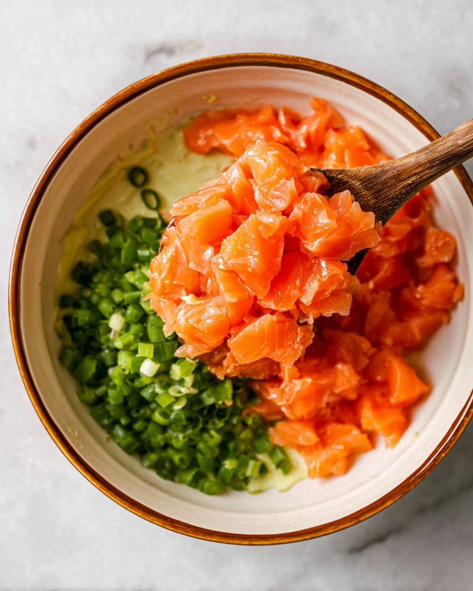 A close-up image shows a white bowl with a brown rim on a white marbled surface. Inside the bowl, there are three visible layers: at the bottom left, finely chopped green scallions; above and centered, a pile of small, bright orange salmon pieces with a fresh and slightly shiny texture; the background inside the bowl has a thin layer of creamy sauce clinging to the sides. A wooden spoon holds a large scoop of the salmon cubes above the scallions, creating a contrast between the green and orange colors. photo taken with an iphone --ar 4:5 --v 7