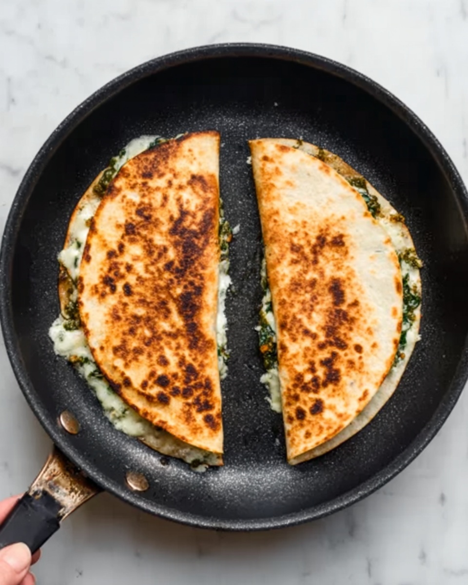 The image shows a black frying pan with two half-circle shaped folded quesadillas inside. Each quesadilla has a golden-brown toasted surface with some darker spots, indicating it was cooked on a hot pan. Inside, melted white cheese and green leafy vegetables are visible where the quesadillas meet in the center. The pan sits on a white marbled surface. A woman's hand is holding the pan handle slightly visible on the left side. photo taken with an iphone --ar 4:5 --v 7