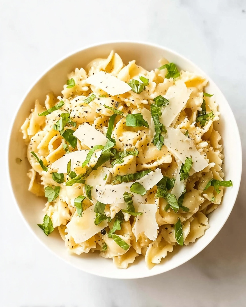 A white bowl is filled with short, twisty pasta that is pale yellow in color. On top, there are scattered thin slices of pale cheese and small pieces of fresh green basil leaves. The pasta looks creamy and slightly shiny, with a sprinkle of black pepper over everything. The bowl sits on a white marbled surface. photo taken with an iphone --ar 4:5 --v 7