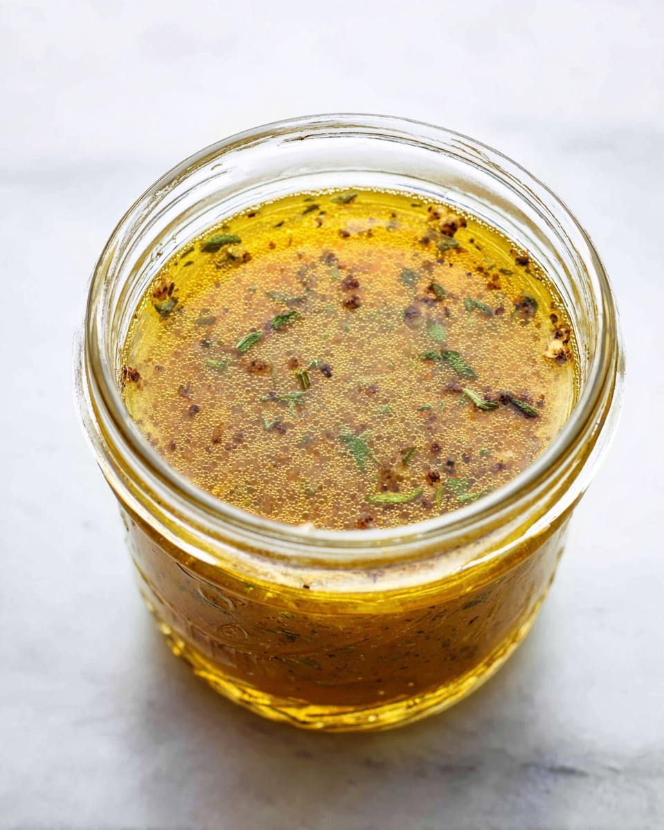 A clear glass jar filled with a three-layer dressing or sauce. The bottom layer is thick and brownish, occupying most of the jar. The top layer is a thin, oily bright yellow layer with visible green and black herbs and spices floating throughout. Tiny bubbles gather on the surface, creating a fresh and textured look. The jar is placed on a white marbled surface. photo taken with an iphone --ar 4:5 --v 7
