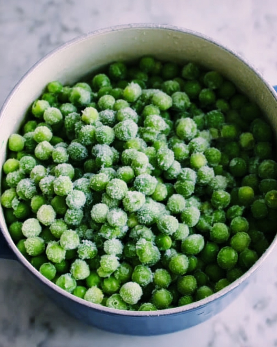 A white pot filled almost to the top with many small round green peas, some with a light frost or ice on them, giving a cold, fresh look. The peas are piled loosely in the pot, showing a bumpy, uneven texture across the surface. The pot sits on a white marbled surface, adding a clean and bright background. Photo taken with an iphone --ar 4:5 --v 7