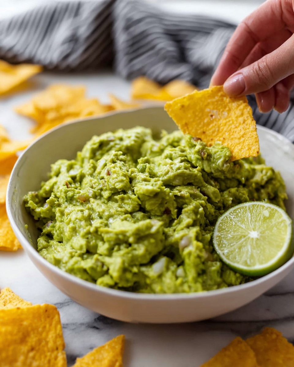 A close-up of a white bowl filled with chunky green guacamole that has a rough texture with visible bits of avocado and small pieces of ingredients mixed in. On the right side of the bowl, a round slice of light green lime is placed on the guacamole. A woman's hand is dipping a triangular yellow corn chip into the guacamole from the left side of the image. The bowl sits on a white marbled surface with extra corn chips scattered in the background and foreground, some blurred. A black and white striped cloth is partly visible behind the bowl. photo taken with an iphone --ar 4:5 --v 7