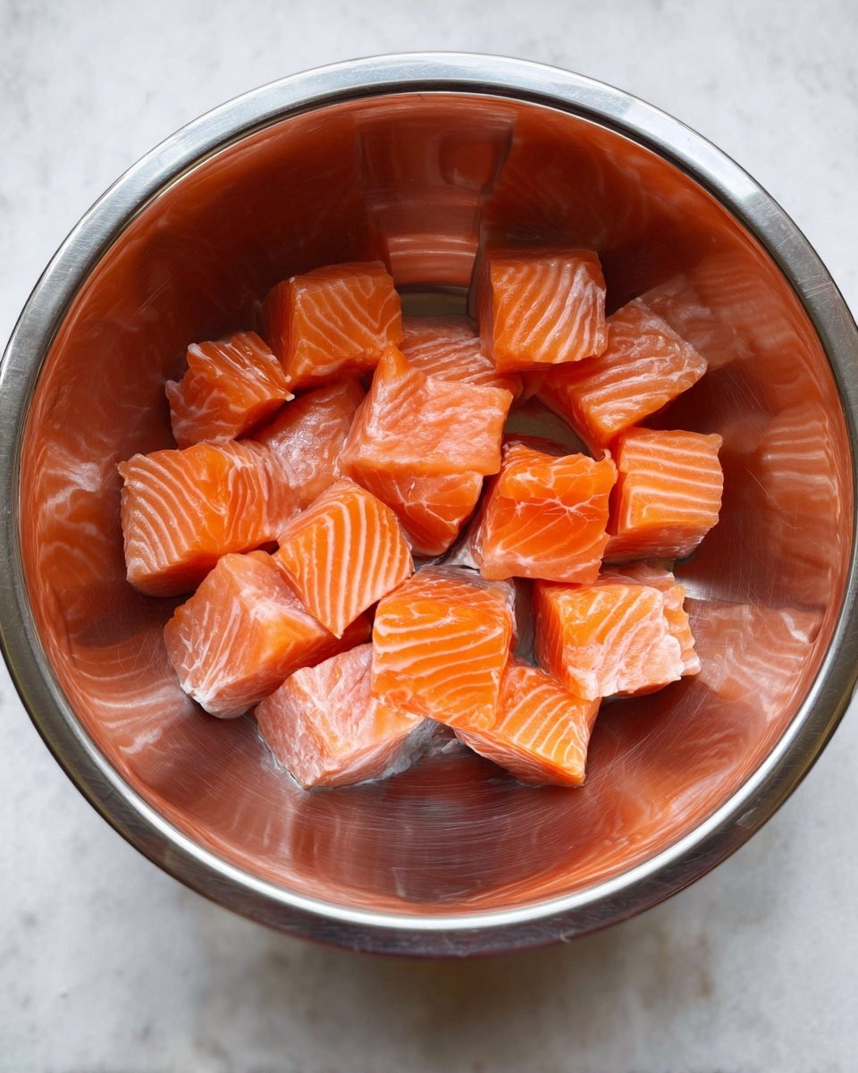 A shiny stainless steel bowl placed on a white marbled surface holds about 15 small cubes of raw salmon. The salmon pieces are bright orange with thin white lines of fat, and some pieces show a bit of pale grey skin attached. The bowl reflects the salmon's color and has smooth, round sides that curve inward, making the fish pieces sit near the center bottom. Photo taken with an iphone --ar 4:5 --v 7