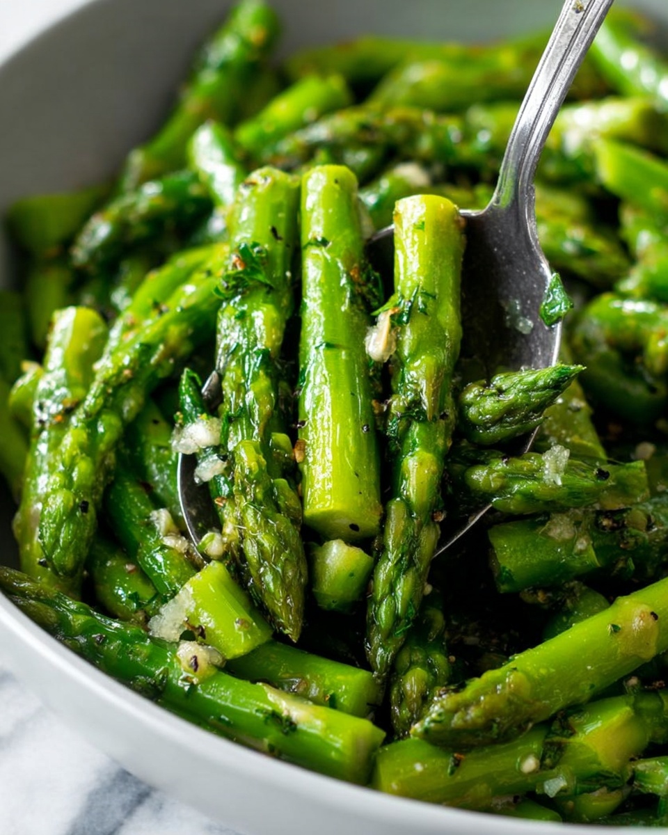 This image shows a close-up of cooked asparagus pieces mixed with small bits of garlic and herbs. The bright green asparagus is cut into roughly equal short sections with visible texture on each spear, some tips are pointed upward. The asparagus is shiny, suggesting it is coated with oil or butter, and small bits of black pepper and herbs scatter across the surface. A silver utensil is scooping the asparagus from a white bowl, which sits on a white marbled surface. Photo taken with an iphone --ar 4:5 --v 7