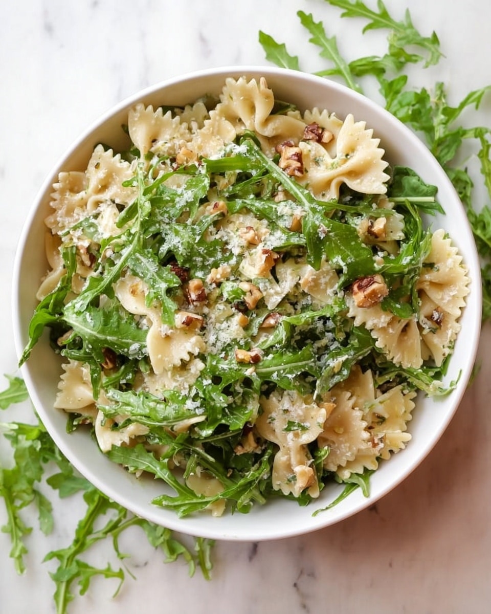 The image shows a white bowl filled with a pasta salad placed on a white marbled surface. Inside the bowl, there are about two layers: a base layer of light cream-colored farfalle pasta with a slightly shiny texture, mixed evenly with fresh, bright green arugula leaves that have a slightly rough texture. Small bits of chopped walnuts and tiny white grated cheese are scattered throughout the salad, adding a speckled effect. Additional arugula leaves are loosely scattered around the bowl on the surface. photo taken with an iphone --ar 4:5 --v 7