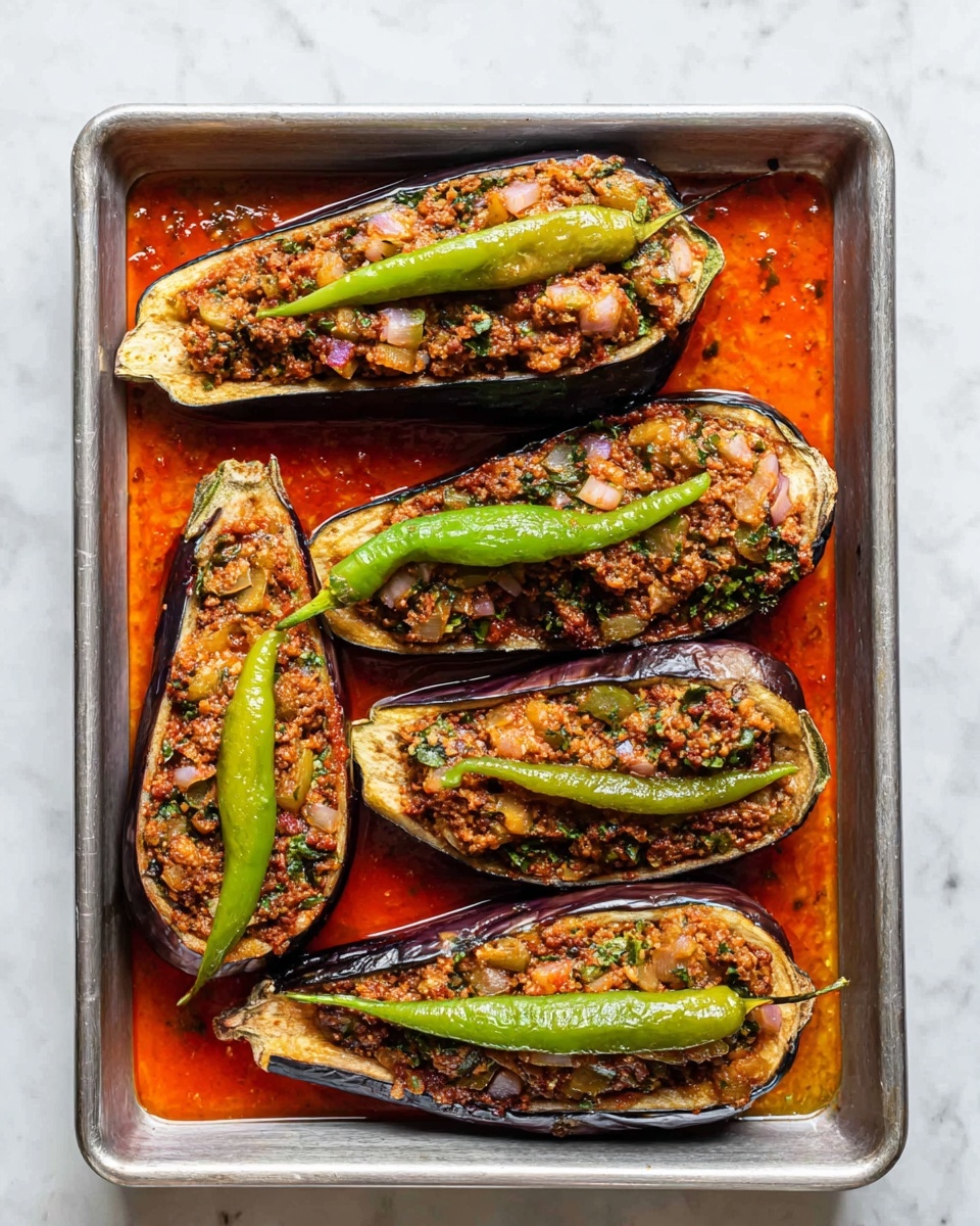 The image shows four halved eggplants placed on a metal baking tray filled with bright red sauce. Each eggplant half has a dark purple skin with a soft, cooked inside filled with a mixed stuffing of light brown minced meat, diced green bell peppers, translucent onions, and bits of herbs, giving a textured and colorful look. On top of the stuffing, there are long, smooth green chili peppers placed neatly on each eggplant, adding a fresh, vibrant contrast to the cooked ingredients. The tray sits on a white marbled surface that adds a clean background to the dish. Photo taken with an iphone --ar 4:5 --v 7