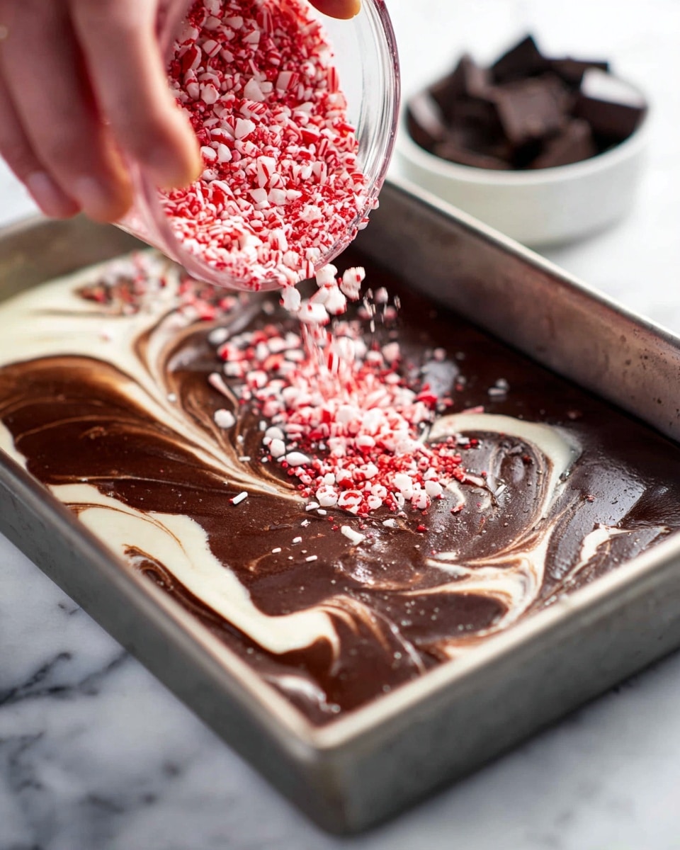 A woman's hand is pouring red and white crushed peppermint sprinkles over a pan filled with a dessert that has two main layers: a thick, creamy white layer at the bottom and a smooth, glossy dark chocolate layer twisted with swirls of white cream on top. The pan is metallic and rectangular, placed on a surface with a white marbled texture. In the background, a small white bowl containing dark chocolate pieces is slightly out of focus. The sprinkles fall unevenly over the dark chocolate layer, adding a festive touch. photo taken with an iphone --ar 4:5 --v 7