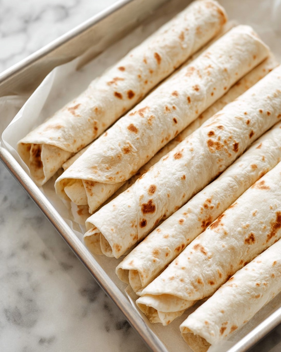 The image shows several rolled tortillas placed closely together on a white tray lined with white paper. Each tortilla is a light beige color with scattered brown spots, showing a soft and slightly textured surface. The rolled tortillas are uniform in size and neatly arranged in rows. The background features a white marbled texture, giving a clean and simple look. photo taken with an iphone --ar 4:5 --v 7