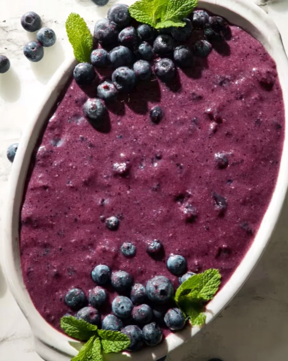 A close-up view of a smooth, thick, purple blueberry smoothie bowl spread evenly inside a white oval dish. The surface of the smoothie has small bits of blueberries visible throughout, giving it a textured look. On the top left corner, there is a cluster of fresh blueberries with bright green mint leaves nearby, and in the bottom right corner, another small pile of blueberries with mint leaves rests against the edge of the bowl. The background is a white marbled surface with soft, natural light creating gentle shadows. Photo taken with an iphone --ar 4:5 --v 7