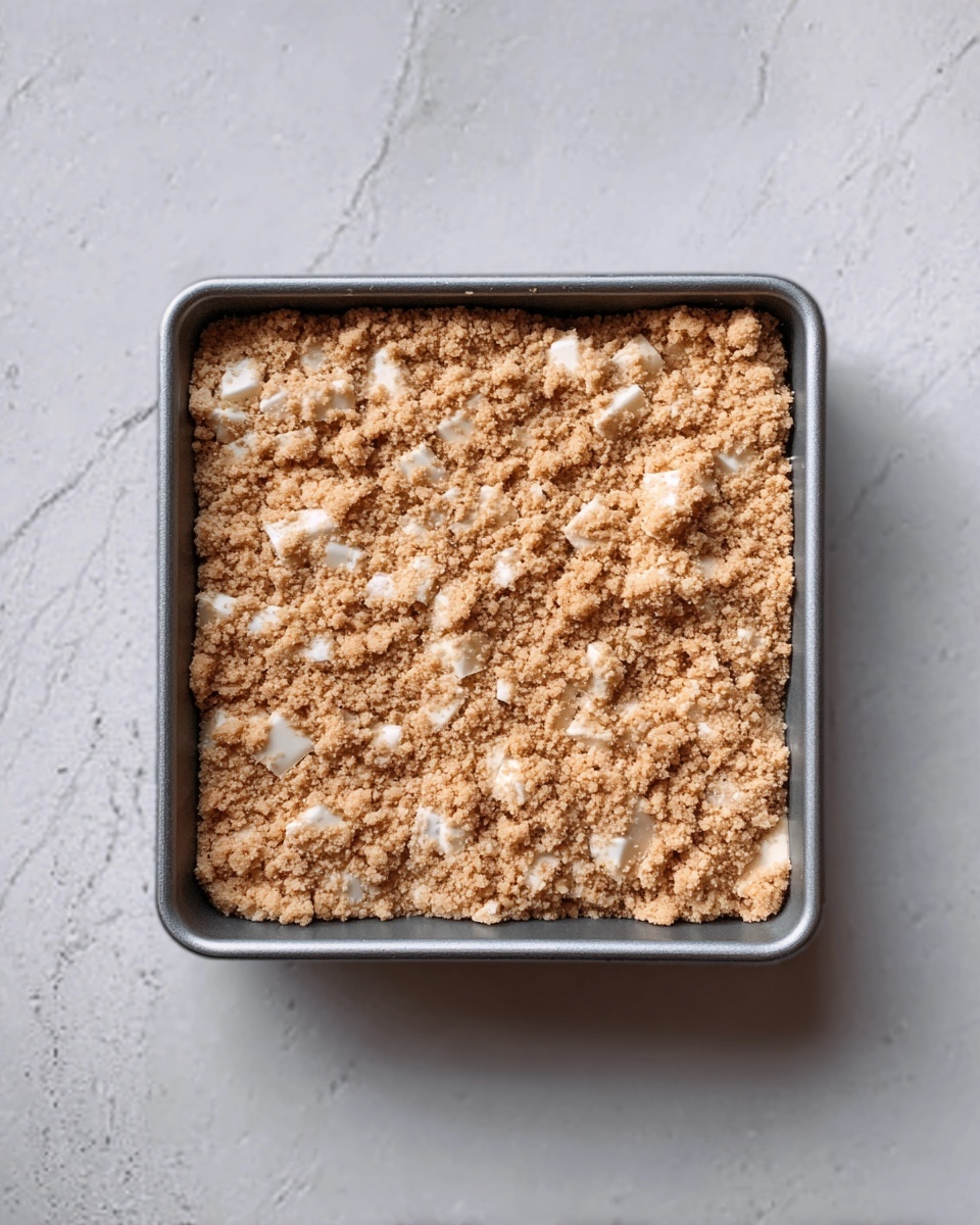 The image shows a square metal baking pan filled with a crumbly topping that looks light brown and golden. The topping is uneven and has white chunks or pieces scattered throughout. The pan is placed on a white marbled surface. The texture of the topping appears crunchy and slightly rough. Photo taken with an iphone --ar 4:5 --v 7