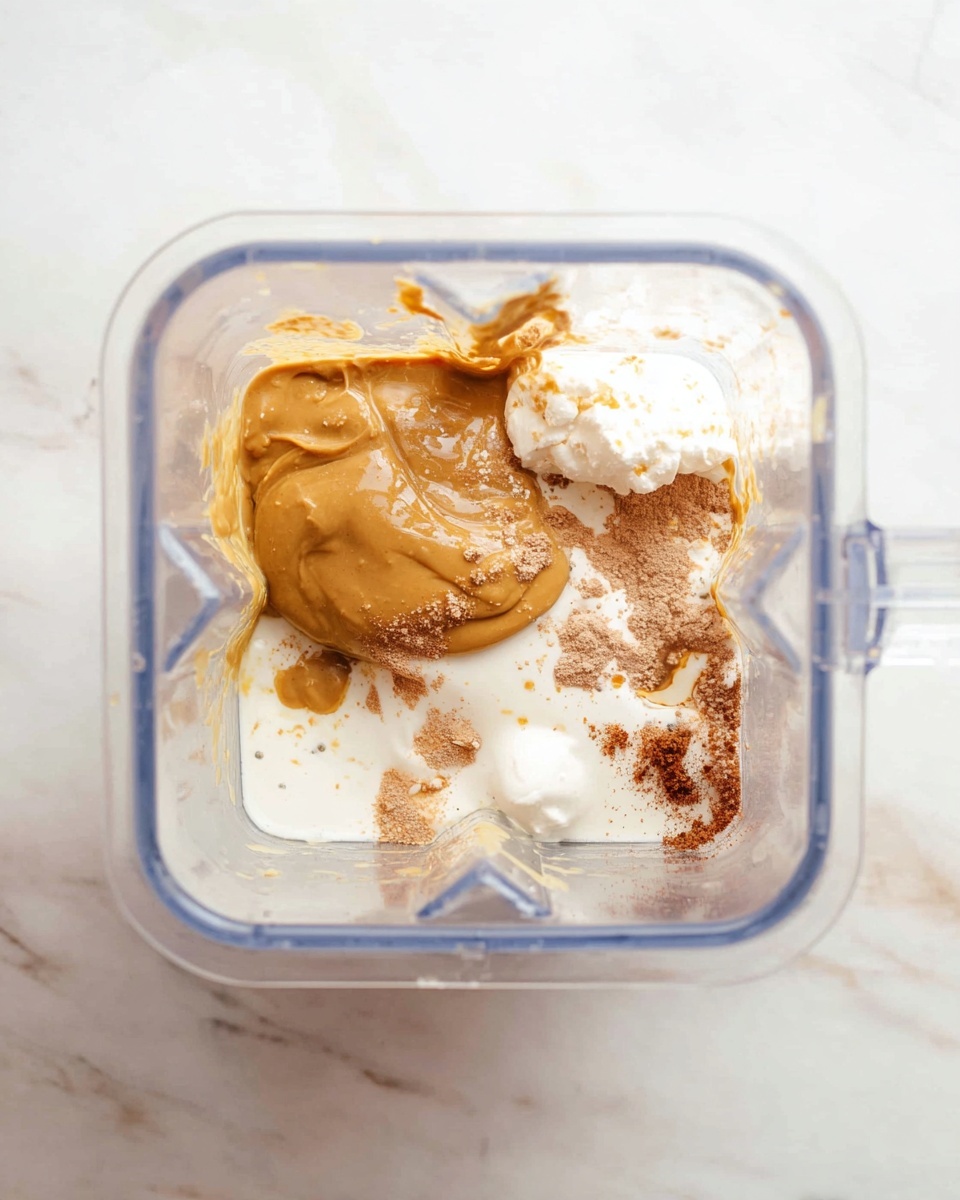Inside a clear square blender container, layers of ingredients rest before mixing. At the bottom layer, there is white liquid with a smooth texture, covering the base. On top of it lies a thick, golden brown dollop unevenly spread with a glossy surface, and near it are white spoonfuls contrasting sharply. Brown specks and powder are scattered around, some blending with the white and golden layers, adding texture. The blender’s transparent walls reflect a bit of the mixture, enhancing the color contrast. The container sits on a white marbled surface. photo taken with an iphone --ar 4:5 --v 7