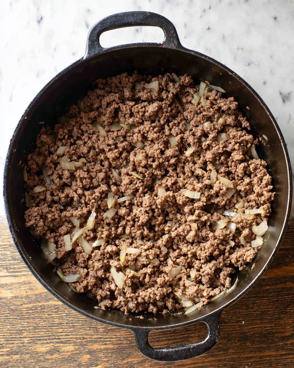 The image shows a black cast iron skillet filled with cooked ground beef that is golden brown and crumbly with small bits of cooked onions or spices mixed in. A wooden spoon is resting in the skillet, scooping some of the beef, with the handle extending out of the skillet. The skillet is placed on a white marbled surface. The lighting is natural and highlights the texture of the meat and the smooth wood of the spoon. Photo taken with an iphone --ar 4:5 --v 7