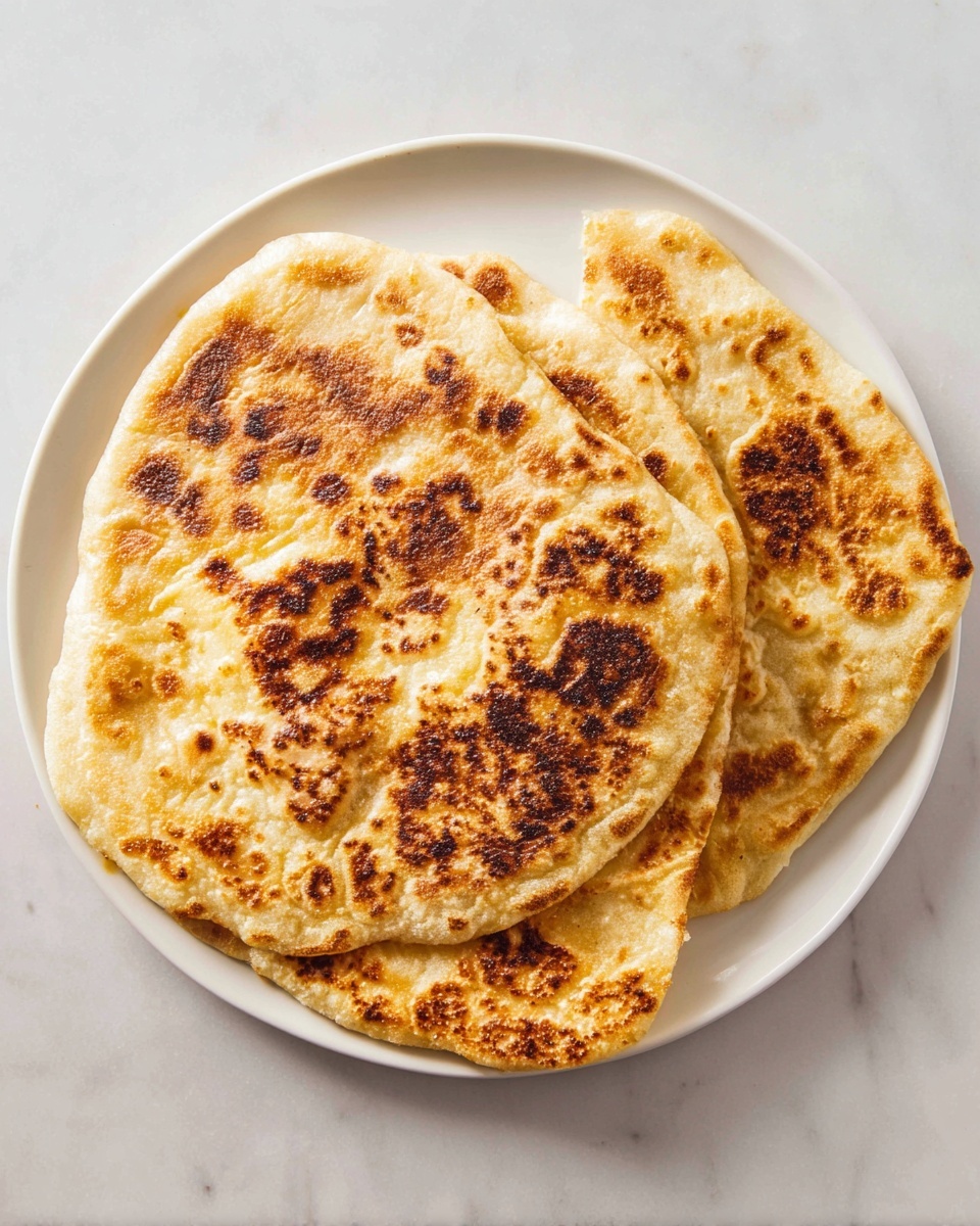 Three pieces of flatbread are layered on a round white plate placed on a white marbled surface. The flatbread has a golden-brown color with darker toasted spots unevenly spread across the surface, showing a slightly crispy and textured appearance. The top flatbread is the smallest and is placed overlapping two larger flatbreads beneath it, creating a layered effect. The edges are irregular and slightly puffy, indicating a soft texture inside. The photo taken with an iphone --ar 4:5 --v 7