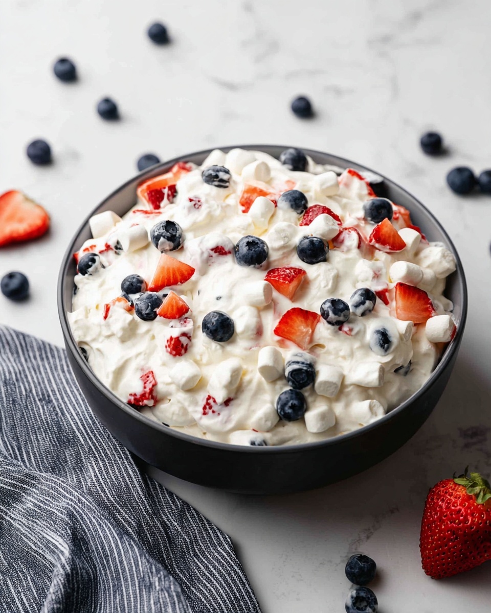 A large dark bowl filled with a thick, creamy white mixture that forms the base layer. Mixed within this creamy layer are small white marshmallows and vibrant chunks of red strawberries alongside whole, dark blue blueberries scattered evenly throughout. The bowl sits on a white marbled surface with scattered blueberries and slices of strawberries around it. A gray striped cloth is partially under the bowl at the front left side, and a whole strawberry is placed to the right of the bowl. The background is a plain white wall. Photo taken with an iphone --ar 4:5 --v 7