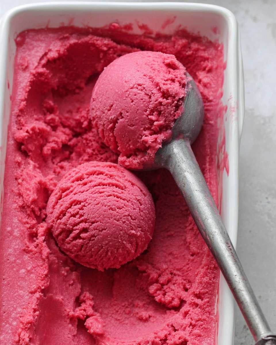 Inside a white rectangular dish, there are two round scoops of bright pink ice cream with a textured surface. The ice cream fills the bottom of the dish in an uneven layer with some splashes on the sides. A silver ice cream scoop with a metal handle is partially buried in the ice cream, holding up one scoop. The background is a soft white marbled texture and the lighting is natural, highlighting the creamy and dense look of the ice cream photo taken with an iphone --ar 4:5 --v 7