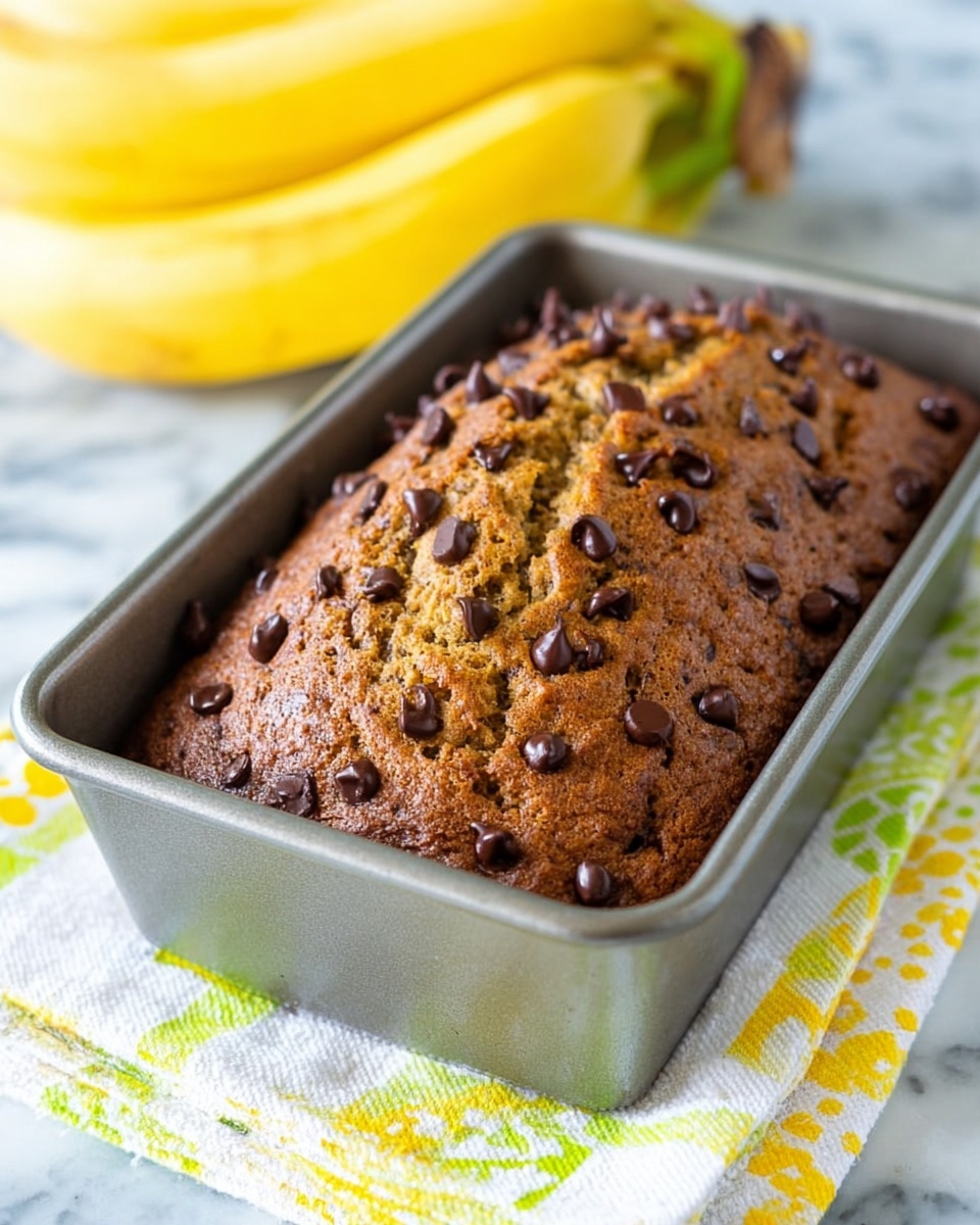 A golden brown banana bread loaf with a textured and slightly cracked top is shown in a metal baking pan. The surface is covered with many small, dark chocolate chips spread evenly. The baking pan sits on a folded cloth with white, yellow, and green patterns. In the background, there is a bunch of ripe bananas with bright yellow skin. The entire scene is set on a white marbled surface. Photo taken with an iphone --ar 4:5 --v 7