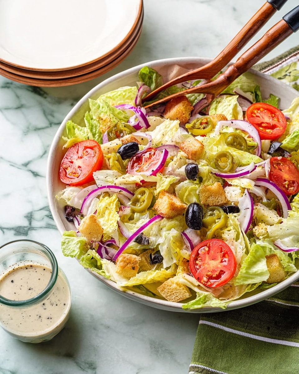 A large white bowl filled with a colorful salad sits on a white marbled surface with a green striped cloth underneath. The salad has a base layer of crisp, light green and pale yellow lettuce leaves, topped with thin rings of purple onion scattered evenly. Bright red tomato slices with visible seeds are placed on top, along with black olives that add dark contrast. Light green pepperoncini peppers add a curved shape, and crunchy, small golden croutons are also sprinkled throughout. Wooden salad tongs with black handles rest inside the bowl, one holding part of the salad. Nearby, a small clear glass holds a creamy dressing with specks of black pepper. A stack of two white plates is seen in the background. photo taken with an iphone --ar 4:5 --v 7