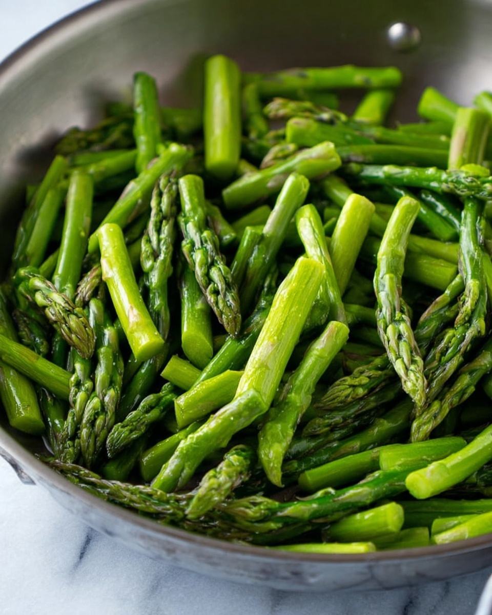 Inside a silver pan, many bright green asparagus pieces are shown, some with pointed tops and others as straight stalks. The asparagus is fresh and has a shiny texture, filling the pan fully and layered with stalks close together and overlapping in different directions. The pan sits on a white marbled surface. The light in the image highlights the fresh green color of the asparagus, reflecting off the smooth pan and the crisp vegetable. Photo taken with an iphone --ar 4:5 --v 7