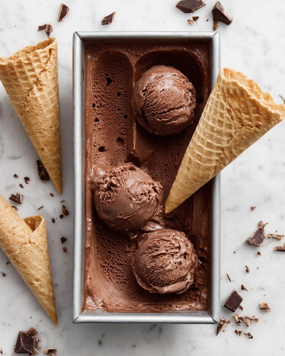 A rectangular metal container filled with rich, dark brown chocolate ice cream, with three visible scoops—one placed near the top left corner, one near the bottom center, and one resting on a waffle cone near the bottom right corner. Another empty waffle cone lies horizontally inside the container near the top right corner. The container is placed on a white marbled surface, scattered with small pieces of chopped chocolate and waffle cone crumbs. Two more empty waffle cones are partially visible, one near the bottom left corner and the other near the left edge. photo taken with an iphone --ar 4:5 --v 7