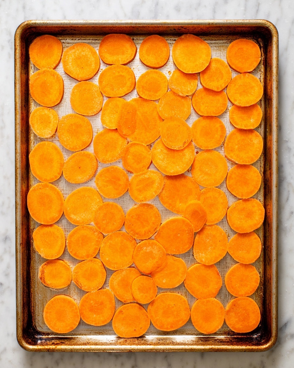 The image shows a metal baking tray placed on a white marbled surface, filled with a single layer of round, thinly sliced bright orange sweet potato pieces spread evenly across the tray. The orange slices vary in size, some larger and some smaller, with a smooth texture and slight earthy marks. There are five rows of slices, creating a neat pattern without overlapping. The tray has a slightly worn look with some browning on the edges, adding a rustic feel. Photo taken with an iphone --ar 4:5 --v 7