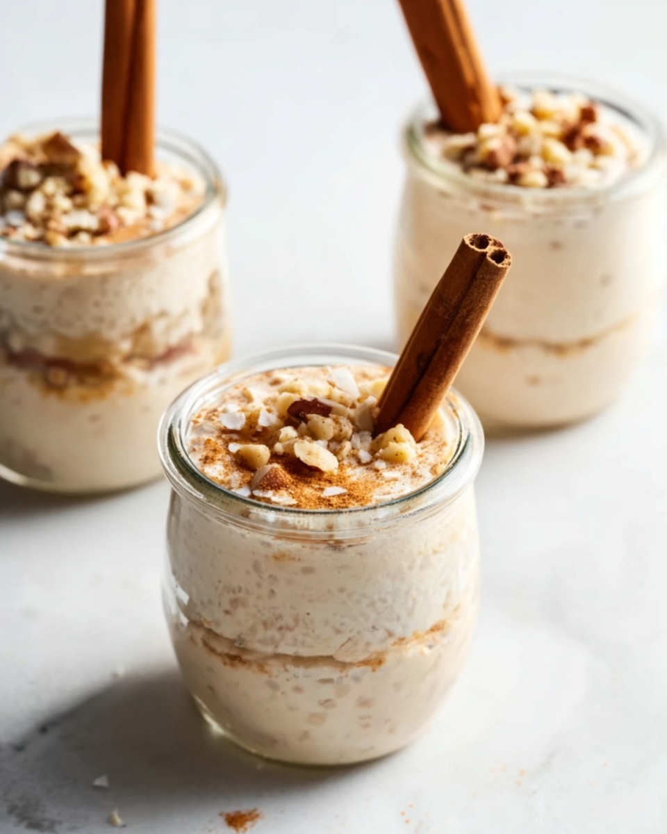 Three small clear glass jars filled with creamy light beige layered dessert are shown on a white marbled surface. Each jar has a cinnamon stick standing upright in the center. The top layer is smooth and creamy with bits of crushed nuts sprinkled over it, adding a rough texture. The jars are placed in a loose triangle shape with one in the front and two blurred in the back. The lighting is soft and natural, highlighting the creamy texture and cinnamon sticks. Photo taken with an iphone --ar 4:5 --v 7