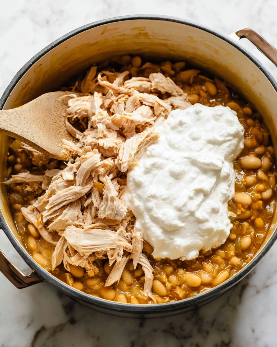 A round white pot filled with three visible layers: the bottom layer is a chunky mixture of yellow beans in a light brown sauce, the middle layer shows shredded light brown chicken pieces spread unevenly on top of the beans, and the top layer is a large dollop of thick, white creamy sour cream centered over the chicken. A wooden spoon with a light brown handle is partially inside the pot, resting diagonally on the left side. The pot sits on a white marbled surface. photo taken with an iphone --ar 4:5 --v 7