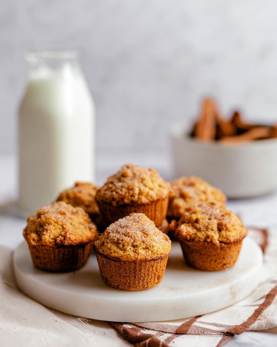 The image shows two pumpkin muffins stacked on top of each other placed on a white speckled plate; the bottom muffin is whole with a golden brown texture while the top muffin is broken in half, revealing a moist orange inside with a crumbly cinnamon sugar topping. Next to the muffins are two cinnamon sticks lying on the plate. The plate is set on another upside-down matching white speckled plate creating a small pedestal. The background features blurred elements like a white bottle, cinnamon sticks in a neutral-colored cup, and a white pumpkin, all on a white marbled surface. Photo taken with an iphone --ar 4:5 --v 7