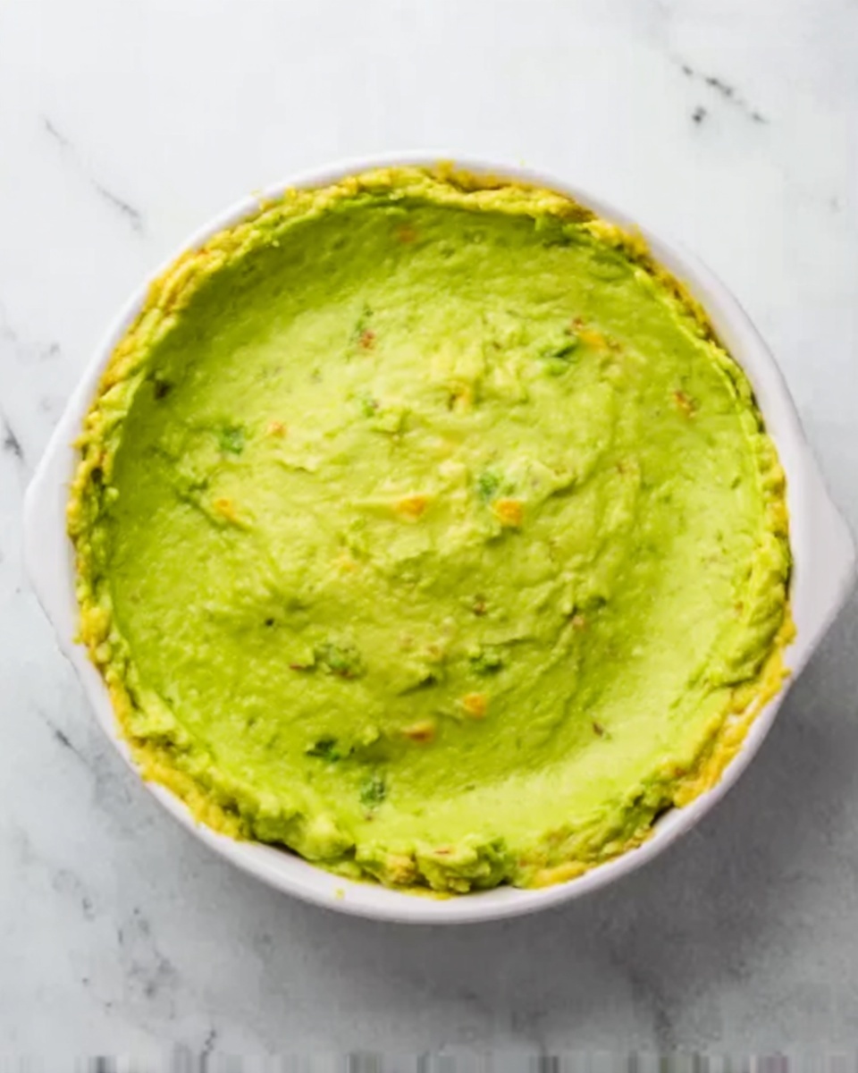 A close-up top view of a white round dish filled with a single thick layer of smooth green mashed avocado spread evenly across the surface. The texture of the avocado shows small bits and a creamy consistency with hints of yellow mixed in. The dish is placed on a white marbled surface. Photo taken with an iphone --ar 4:5 --v 7