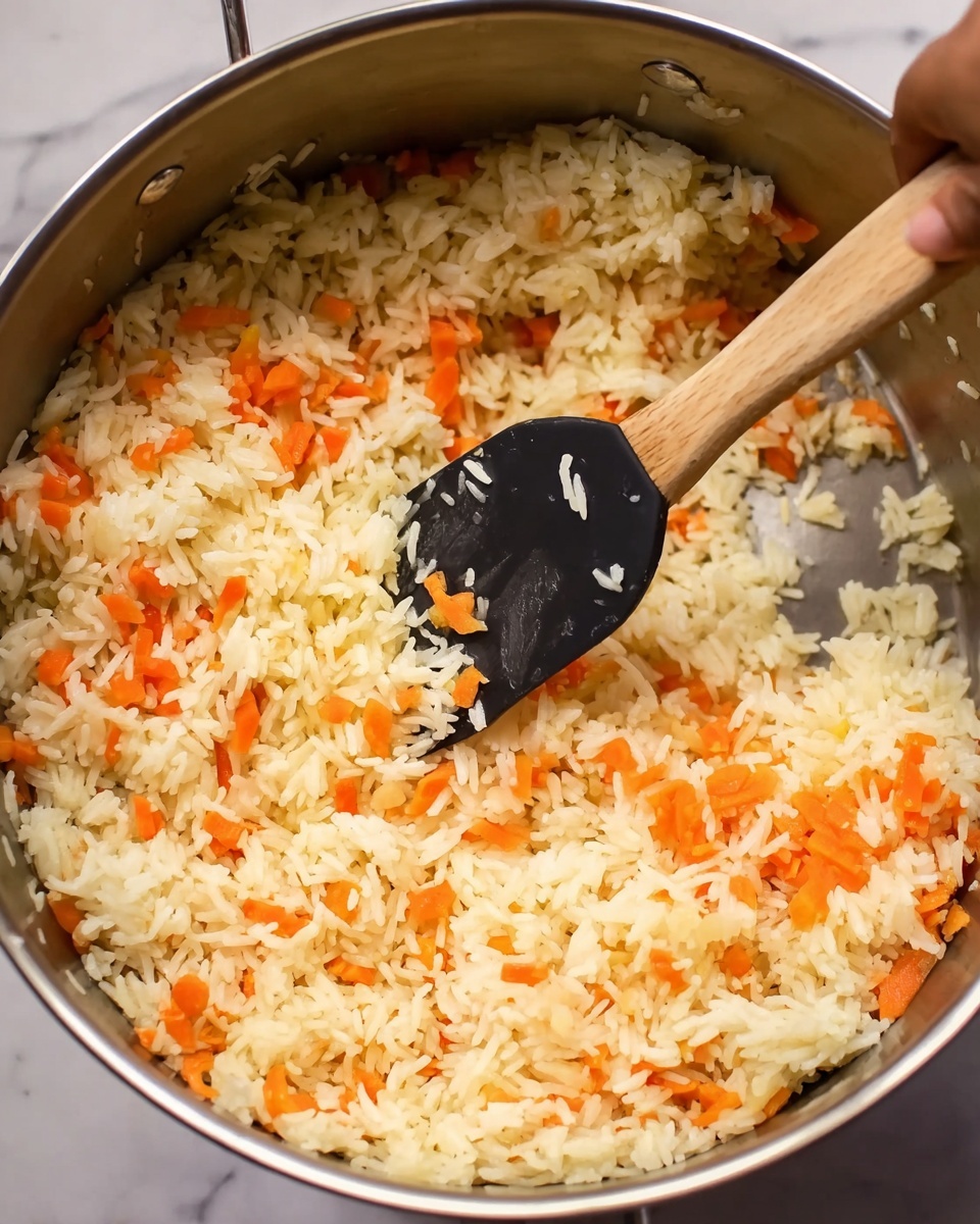 This image shows a close-up of cooked white rice mixed with small orange carrot slices inside a shiny silver pot. The texture of the rice is soft and fluffy, while the diced carrot pieces add bright orange color throughout. A black spatula with a wooden handle is stirring the rice and carrots, and a woman's hand is holding the spatula. The shiny metal pot reflects light softly, set against a white marbled surface. photo taken with an iphone --ar 4:5 --v 7