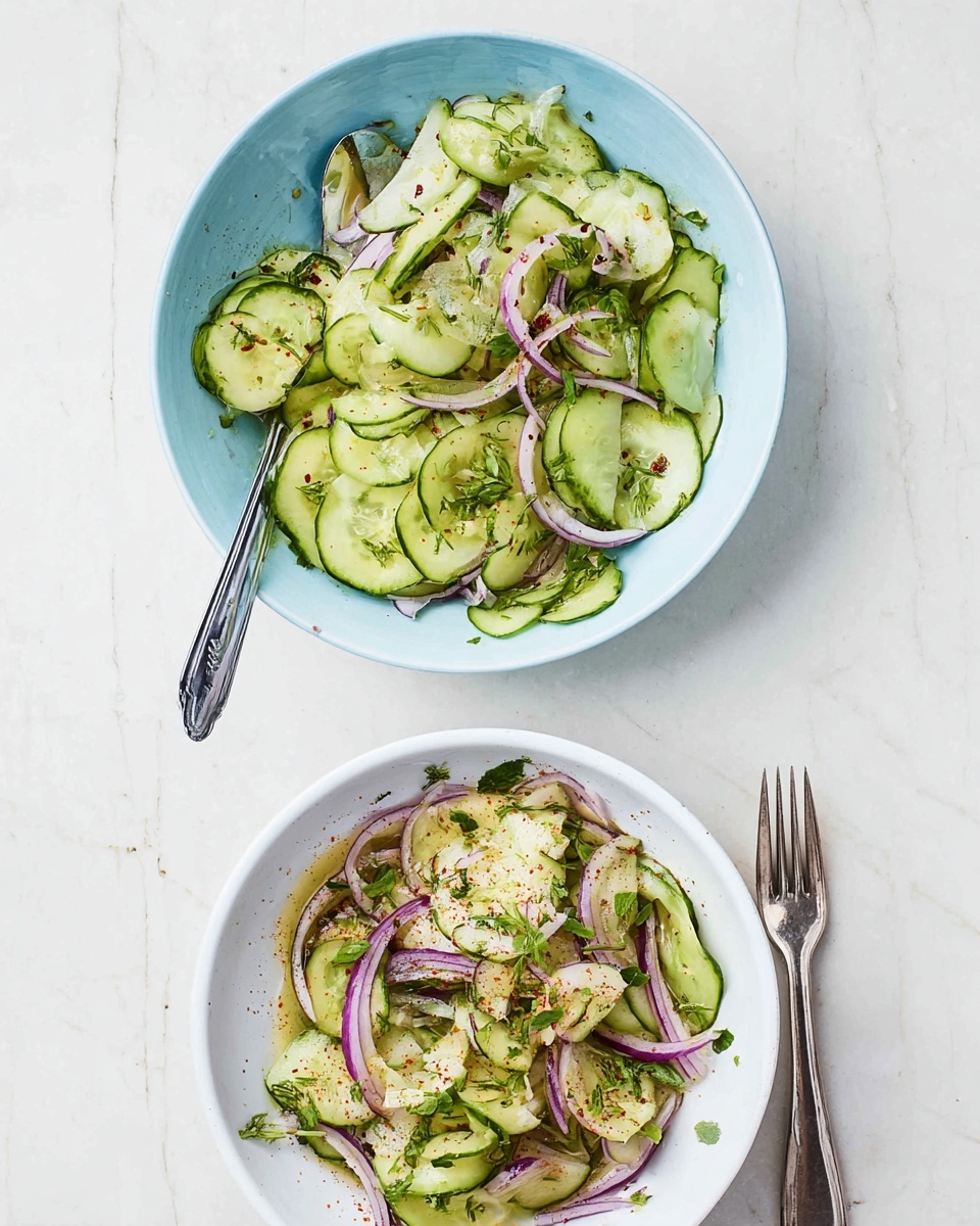 Two bowls of cucumber salad are placed on a white marbled surface. The bowl at the top is light blue and filled with thin sliced pale green cucumbers mixed with thin slices of light purple onions and small green herbs scattered throughout. A silver spoon is resting inside this bowl. The bowl at the bottom is white and also filled with thin sliced cucumbers layered with thin purple onion slices, sprinkled with green herbs, and a light dusting of red spices on top. A silver fork lies beside this bowl on the right side. The colors are fresh and bright, showing a simple, healthy salad. Photo taken with an iphone --ar 4:5 --v 7