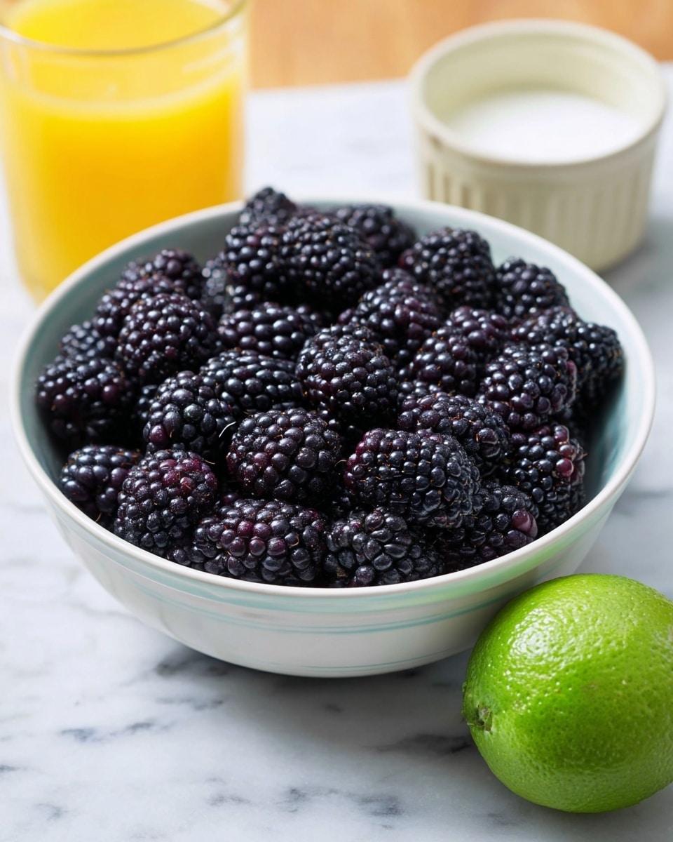 A close-up image of a white bowl filled with many dark purple blackberries, showing their shiny, bumpy texture and rich color. Behind the bowl, there is a clear glass with orange juice and a white bowl with yogurt. A whole fresh lime with a smooth, bright green skin is placed in front of the bowl on a white marbled surface. The overall setting gives a fresh and healthy vibe. photo taken with an iphone --ar 4:5 --v 7