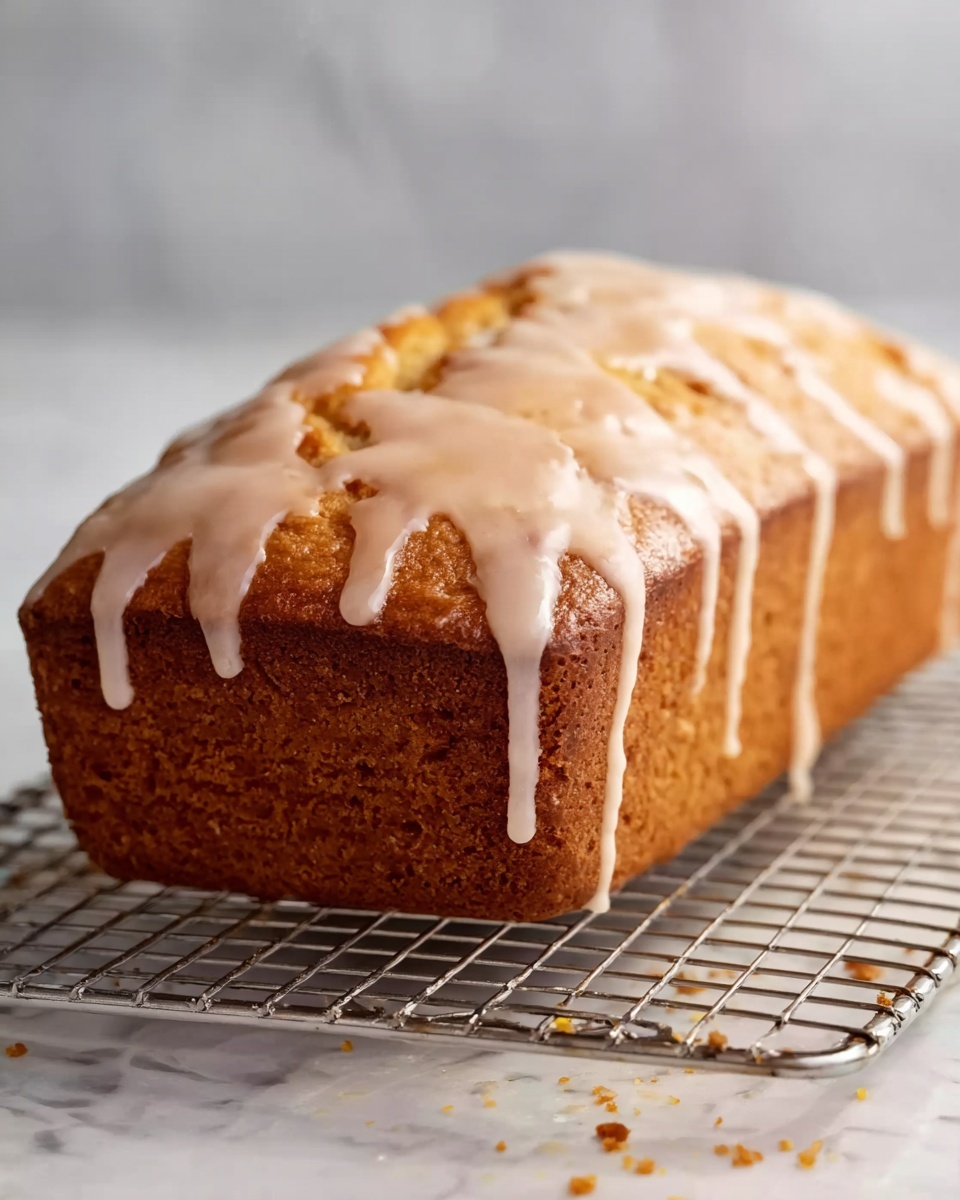 A loaf of golden brown cake with a slightly rough texture on the sides, topped with a smooth, shiny light beige glaze dripping down the edges in uneven lines. The cake rests on a silver wire cooling rack, with some crumbs scattered around. The background is a white marbled surface, softly blurred. Photo taken with an iphone --ar 4:5 --v 7
