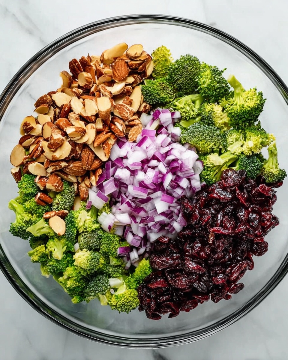 A clear glass bowl on a white marbled surface holds four separate groups of salad ingredients arranged side by side without mixing. The first group is light brown chopped almonds with a rough texture, the second is small bright green broccoli florets, the third group is small purple and white diced red onions, and the last group consists of dark red dried cranberries with a shriveled texture. The ingredients fill the bowl in an even circle. Photo taken with an iphone --ar 4:5 --v 7