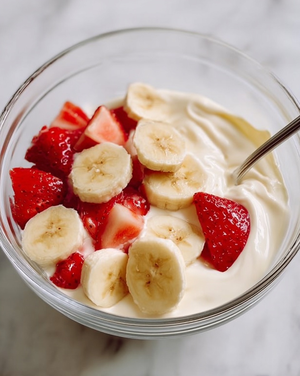A clear glass bowl holds a creamy white yogurt base filling the bottom layer with a smooth texture. On top, there are two layers of fresh fruit: round pale yellow banana slices and bright red strawberry pieces, some triangular and some cut in halves, spread across the yogurt. A silver spoon rests inside the bowl, partially tucked among the fruit pieces. The bowl sits on a white marbled surface. photo taken with an iphone --ar 4:5 --v 7