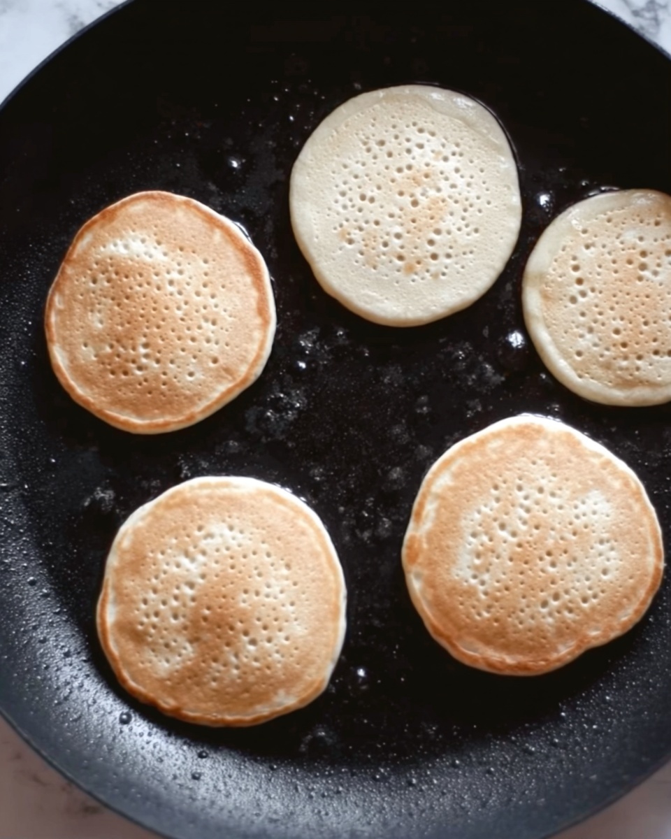 The image shows five small pancakes cooking on a dark pan, each pancake round with small holes appearing on the top surface, signaling they are cooking through. The pancakes have a light beige color with a slightly smooth texture. There is no plate or background visible, only the black pan with small oil spots around the pancakes. The surface beneath the pan has been changed to white marbled texture. photo taken with an iphone --ar 4:5 --v 7