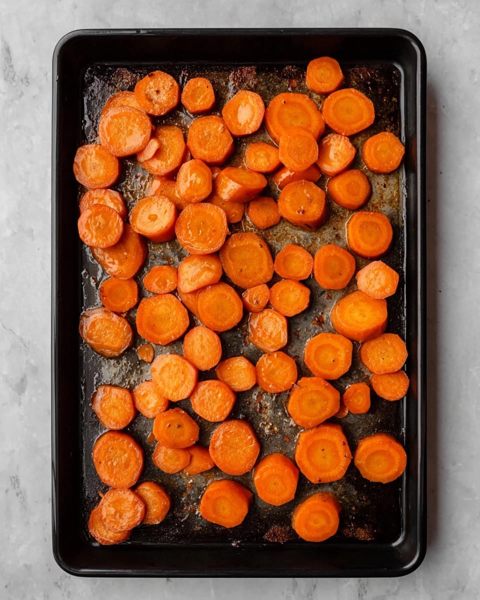 The image shows a black baking tray filled with about 40 pieces of round carrot slices. The carrot slices are bright orange, shiny from oil, and spread out in a single layer, some of them slightly leaning on each other. The tray has some brown spots from roasting, and the whole scene is set on a white marbled texture background. Photo taken with an iphone --ar 4:5 --v 7