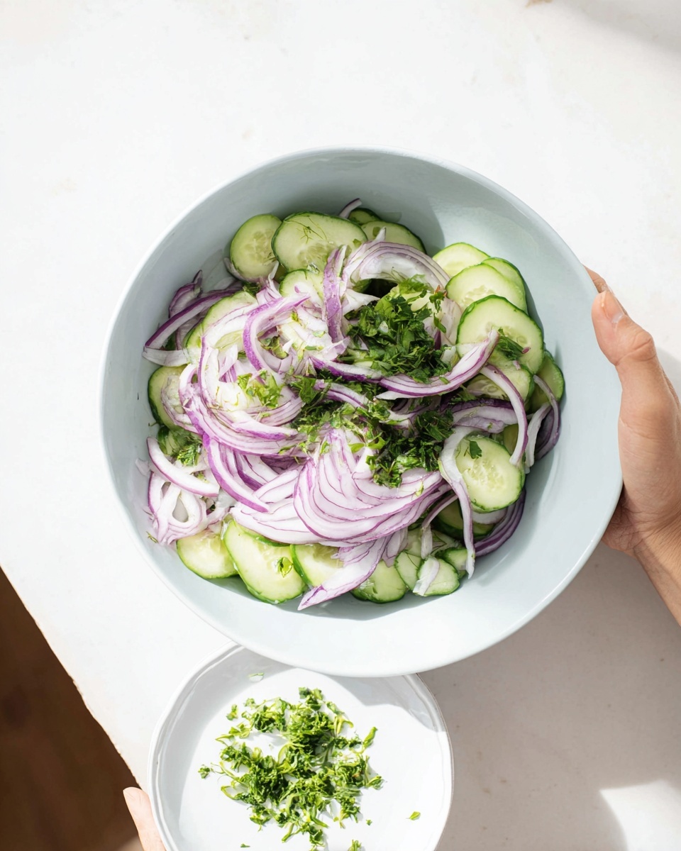 A white bowl holds a fresh salad with three visible layers: the bottom layer is pale green cucumber slices, the middle layer is chopped green herbs, and the top layer is thinly sliced purple-red onion rings spread evenly. A woman's hand holds a small white plate with scattered chopped green herbs near the bottom right corner. The setting is on a white marbled surface with soft natural light. photo taken with an iphone --ar 4:5 --v 7