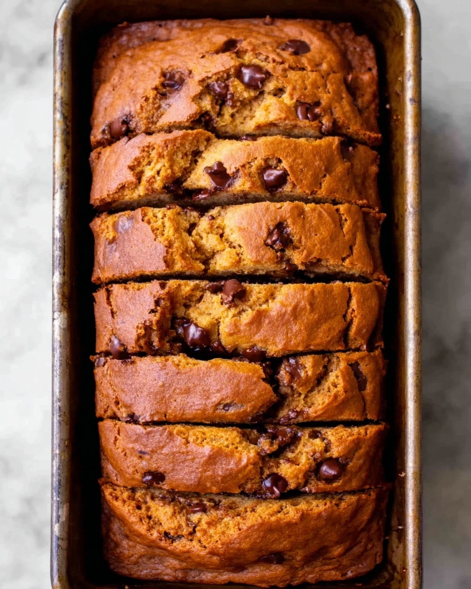 A rectangular loaf of brown chocolate chip bread is shown from above, sliced into about ten even pieces. The bread has a slightly rough, cracked surface with visible dark chocolate chips spread throughout the soft interior. The bread sits in a metal baking pan with a slightly worn look, placed on a white marbled surface. The texture looks moist and dense, with the chocolate chips giving extra detail and richness to the golden brown outer crust photo taken with an iphone --ar 4:5 --v 7