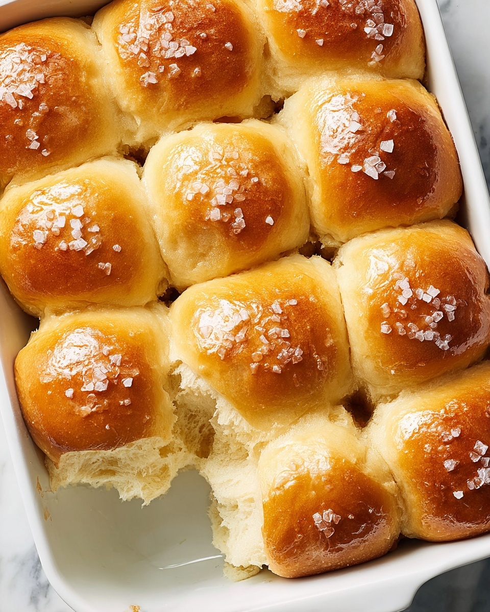 The image shows a close-up of six golden brown soft dinner rolls stacked together on a white plate. Each roll has a shiny, smooth top sprinkled with flakes of coarse salt. The rolls are fluffy and light inside, visible where a piece has been bitten off. The background is a white marbled surface, adding a clean and simple look to the scene. photo taken with an iphone --ar 4:5 --v 7