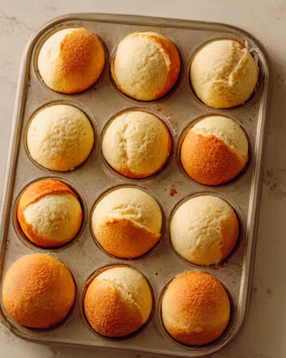 The image shows a metal muffin tray filled with 12 round bread rolls. Each roll is slightly risen and golden brown on top, with some slightly split open showing a soft, light beige inside. The tray is placed on a white marbled surface with warm, natural light creating soft shadows. Some rolls have a smooth texture while others have small cracks and uneven golden hues, giving a fresh baked look. The overall color palette is warm beige and golden brown, highlighting the bread’s texture. Photo taken with an iphone --ar 4:5 --v 7