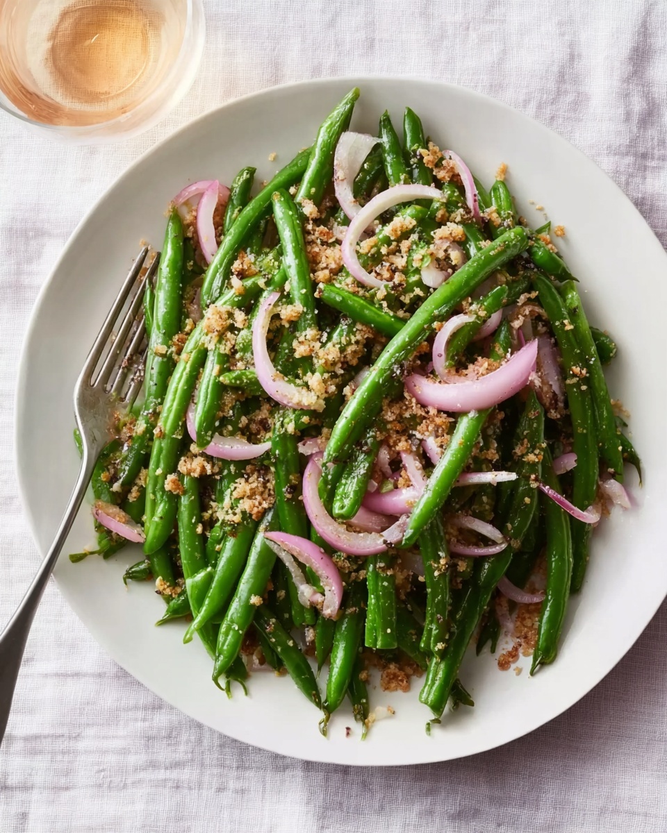 A white plate with a piled layer of bright green cooked green beans as the base, topped with a mix of finely chopped red onions, small green capers, and yellow mustard seeds sprinkled evenly over the beans, adding texture and color contrast. The whole presentation sits on a white marbled surface with a blurred blue cloth in the background. photo taken with an iphone --ar 4:5 --v 7