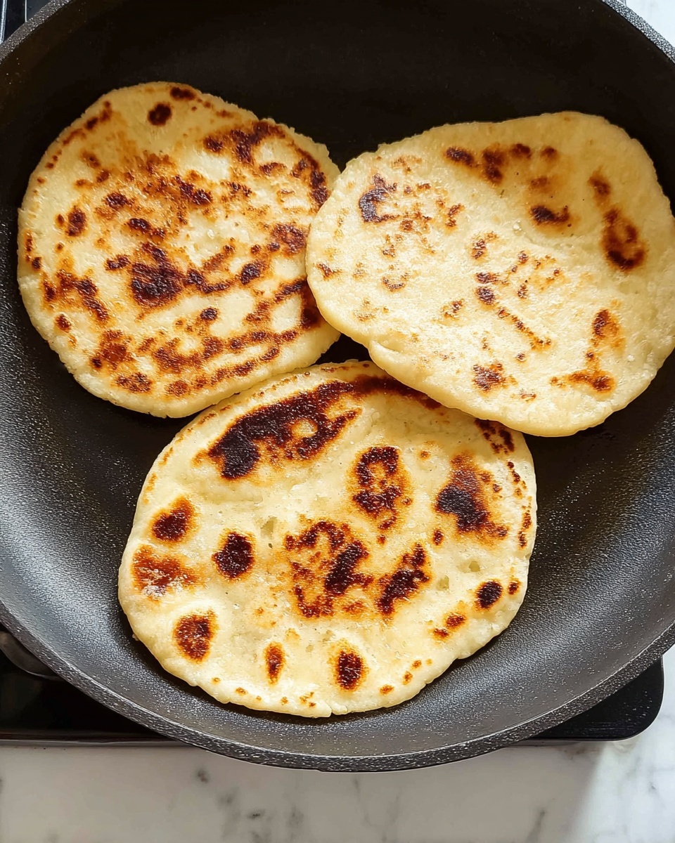 Three round flatbreads with uneven, golden brown spots cook in a black skillet. Each flatbread shows different patterns of toasting with some darker brown burnt patches scattered across the light beige surface. The skillet is placed on a stove with a white marbled background visible below. The flatbreads have a slightly puffy texture and are spaced apart in the pan, presenting a warm, fresh-cooked look photo taken with an iphone --ar 4:5 --v 7