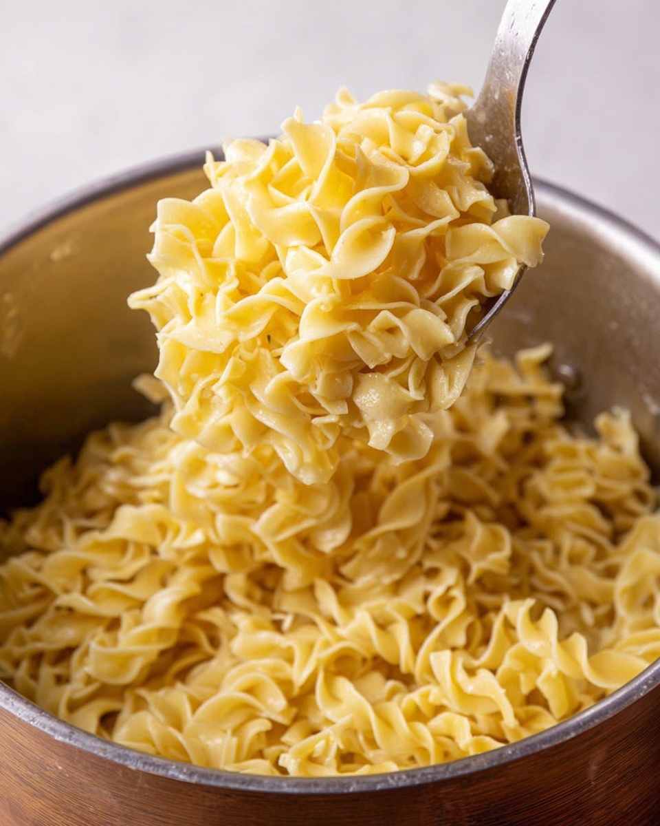 A close-up view of a silver pot filled with soft, yellow egg noodles coated in a light sauce. The noodles are curly and thick, piled high inside the pot, with a large silver spoon lifting a generously filled scoop of the noodles above the pot. The noodles have a slightly shiny texture and appear warm and tender. The background shows a hint of light wood, while the surface beneath the pot is a white marbled texture. photo taken with an iphone --ar 4:5 --v 7